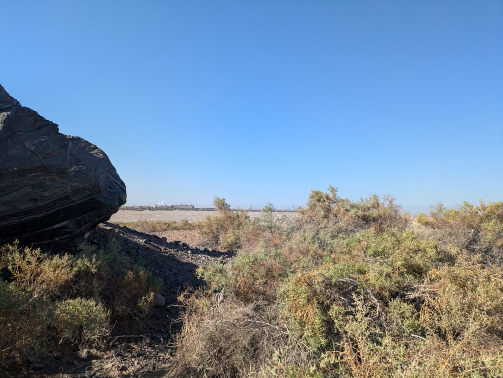 A large dark rock in a desert landscape with an industrial site in the distance